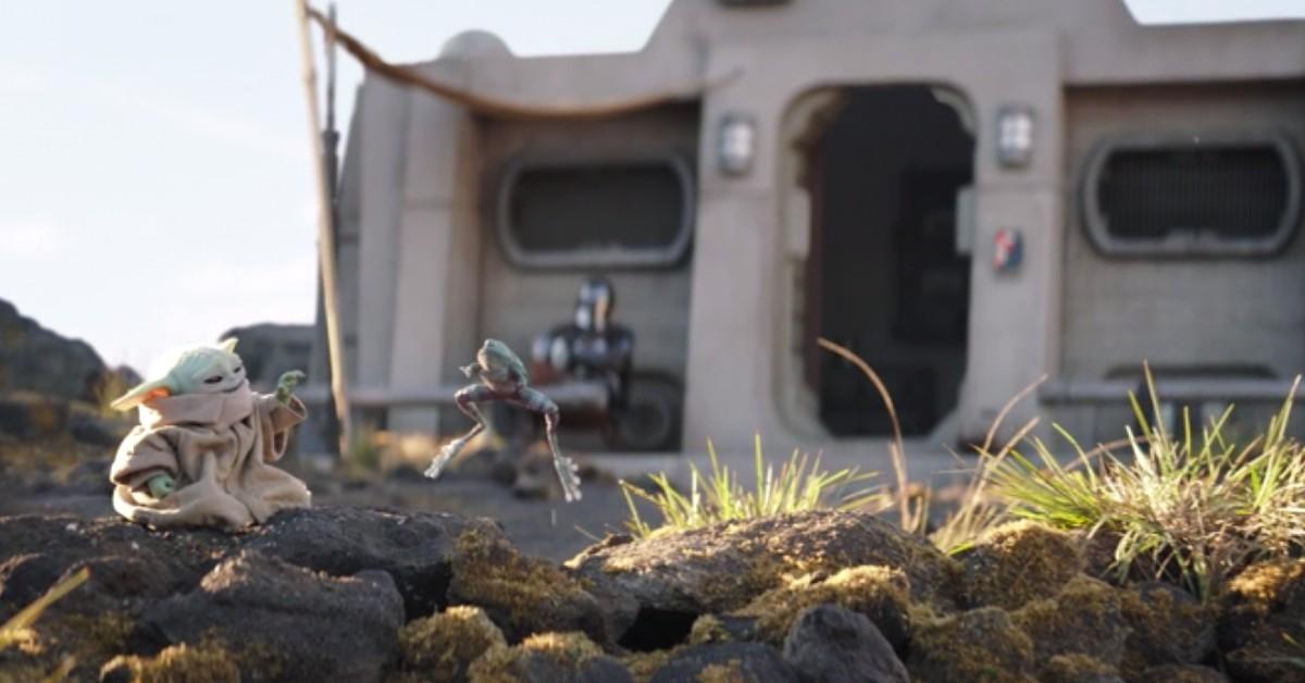 Grogu (L) Force lyfter en groda medan Din tittar på i bakgrunden framför deras stuga i Nevarro. 
