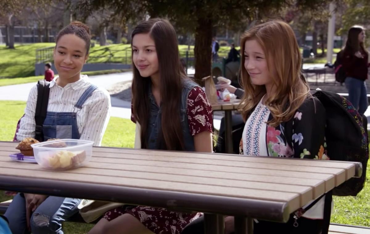 「ニュー・ガール」の学生としてのオリビア・ロドリゴは、他の 2 人の女の子と屋外のピクニック テーブルに座っています。
