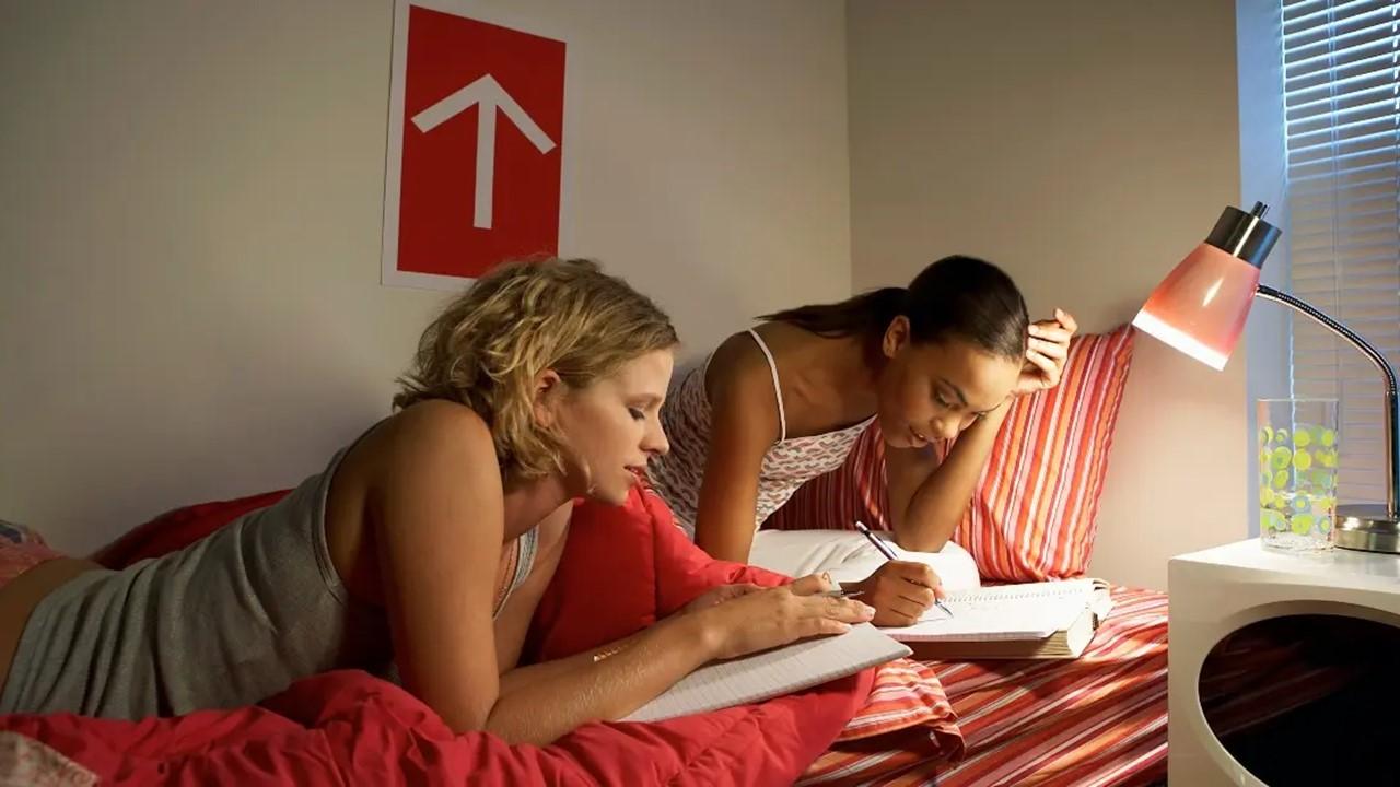 Två unga kvinnor som sitter på sängen och studerar bredvid en lampa