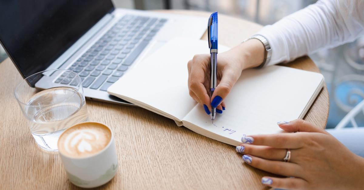 Frau mit lackierten Nägeln schreibt ein Buch in ein Notizbuch, während sie vor einem Laptop sitzt.