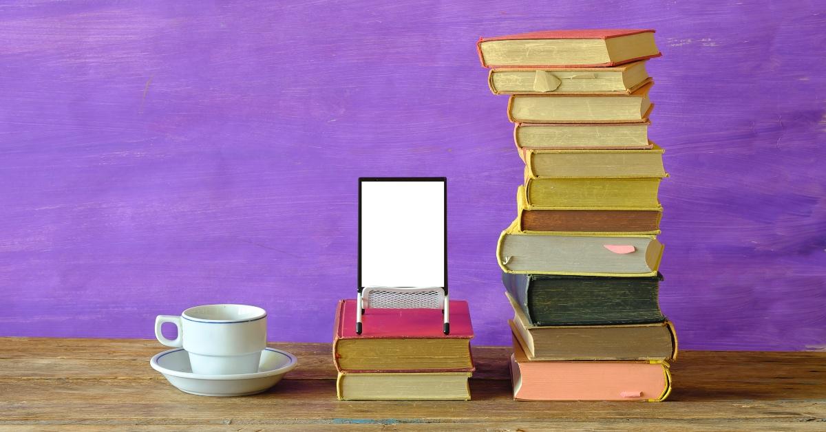 Stak bøger ved siden af ​​en e-læser foran en lilla vægbaggrund. 