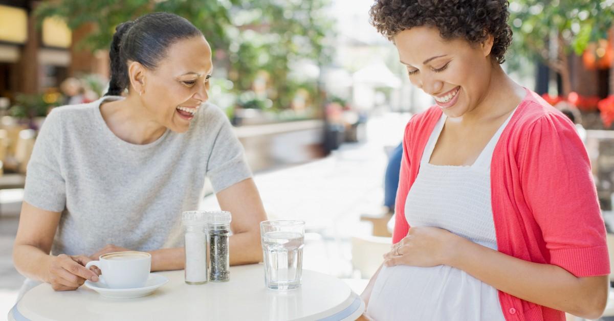 一位朋友和她怀孕的朋友坐在咖啡馆里，后者捧着她的肚子微笑。 