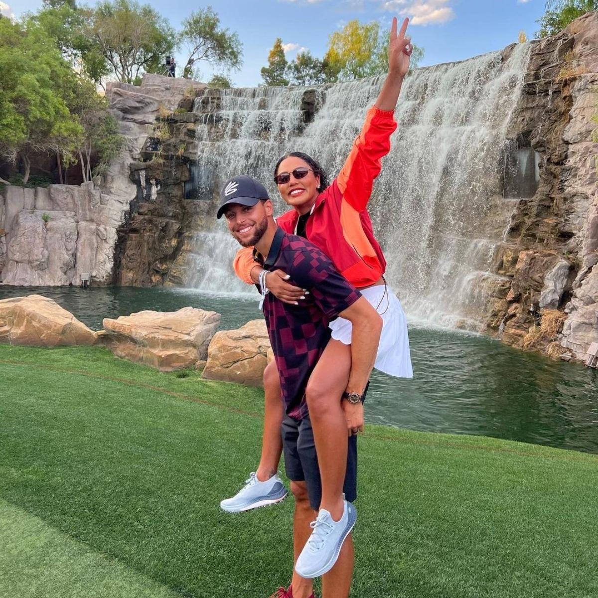Ayesha Curry rider på Steph Currys rygg, ler framför ett vattenfall.