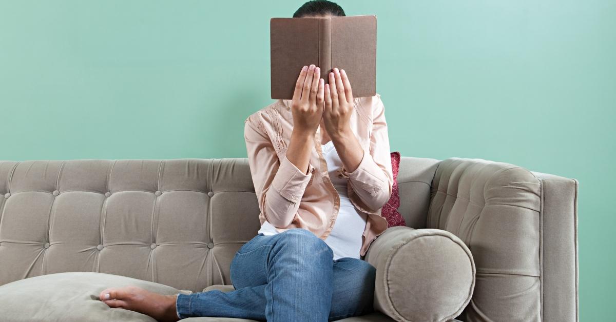 Frau hält Buch auf Couch hoch und bedeckt ihr Gesicht damit