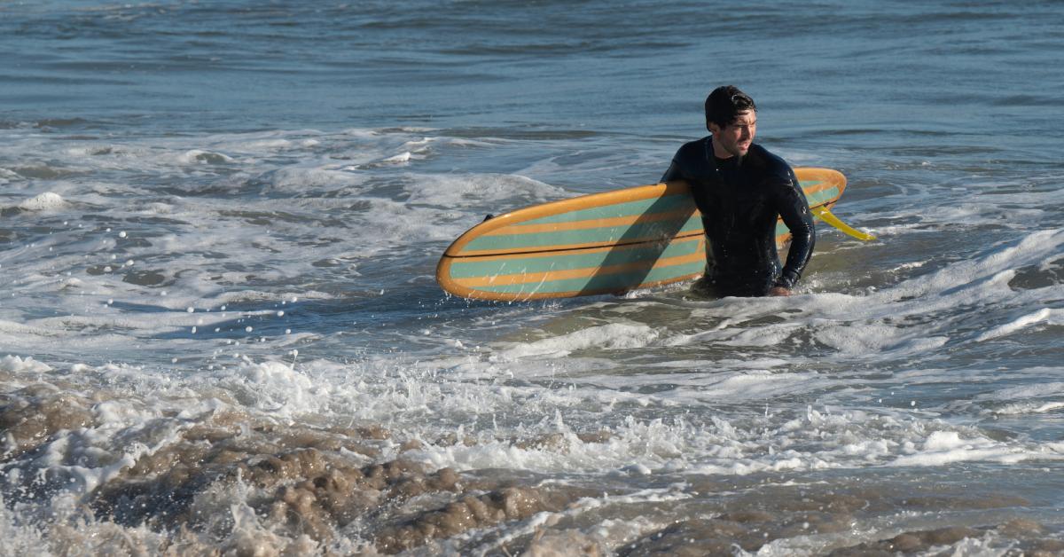 Joey Graziadei, vestito con una muta, guarda di lato mentre emerge con grazia dall'oceano, con la tavola da surf in mano.